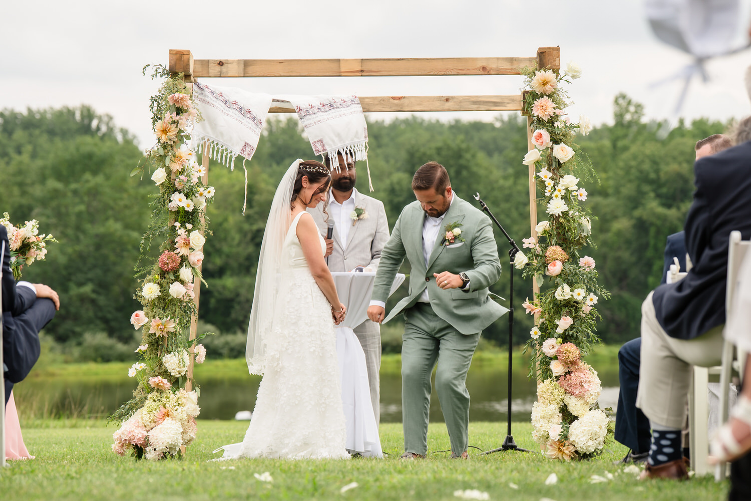 A bride and groom stand under a floral arch outdoors. The groom steps on a glass, performing the Jewish wedding tradition. A person officiates the ceremony. Guests are seated on either side. Trees and greenery are in the background.