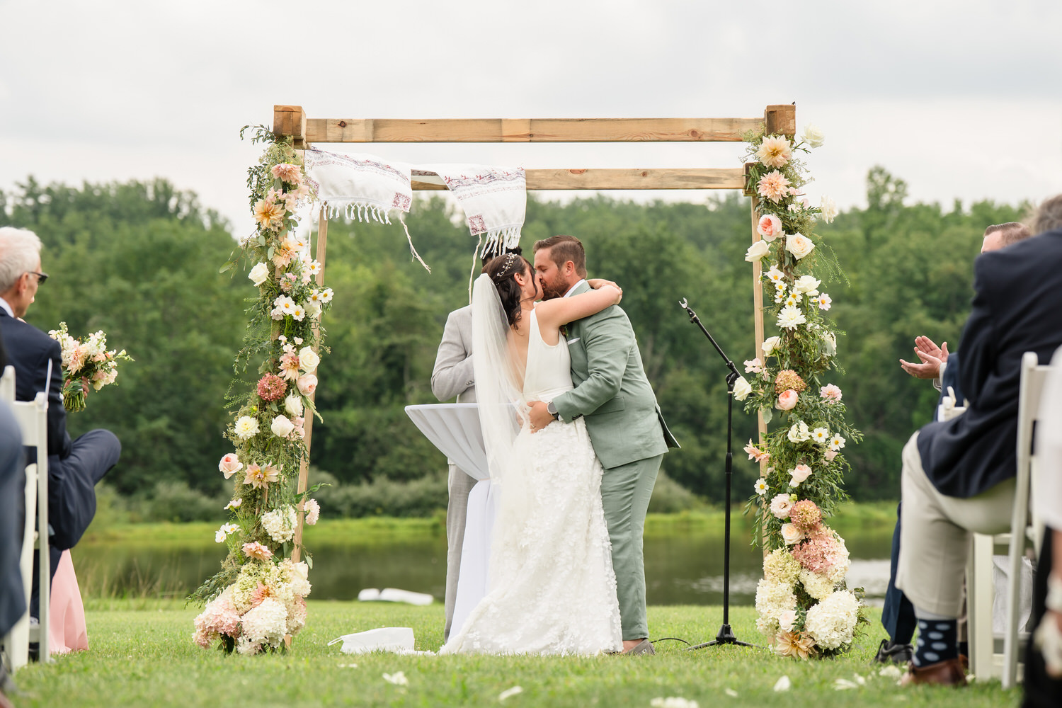 A bride and groom kiss under a flower-adorned wooden arch at an outdoor wedding ceremony, surrounded by seated guests and greenery in the background.