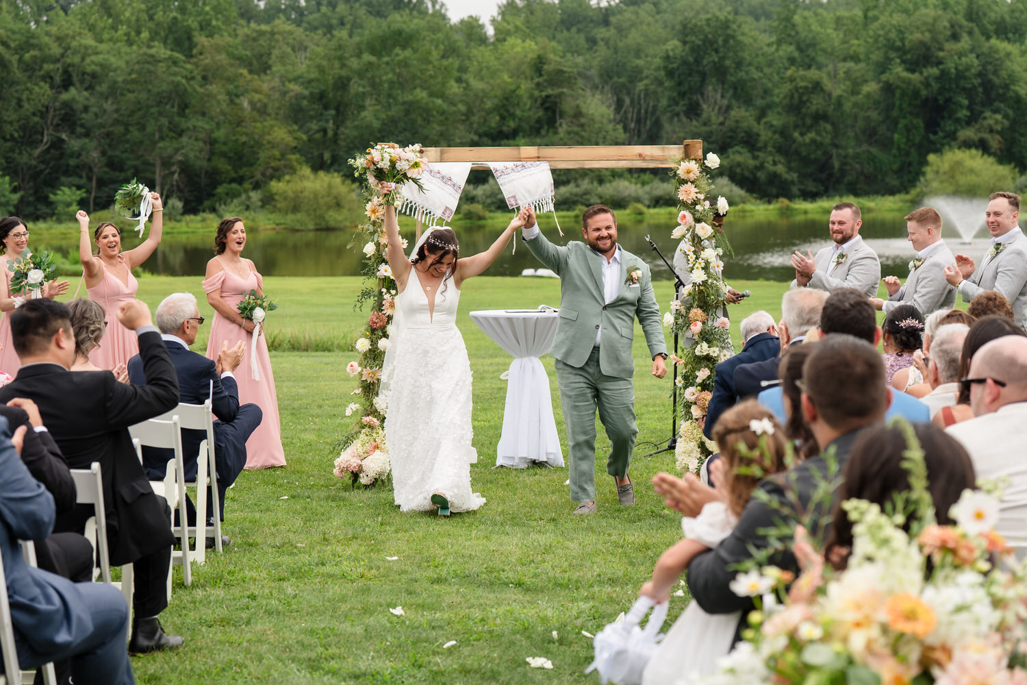 A bride and groom hold hands and celebrate under a floral wedding arch outdoors, surrounded by cheering guests. Bridesmaids in pink dresses and groomsmen in gray suits stand nearby.