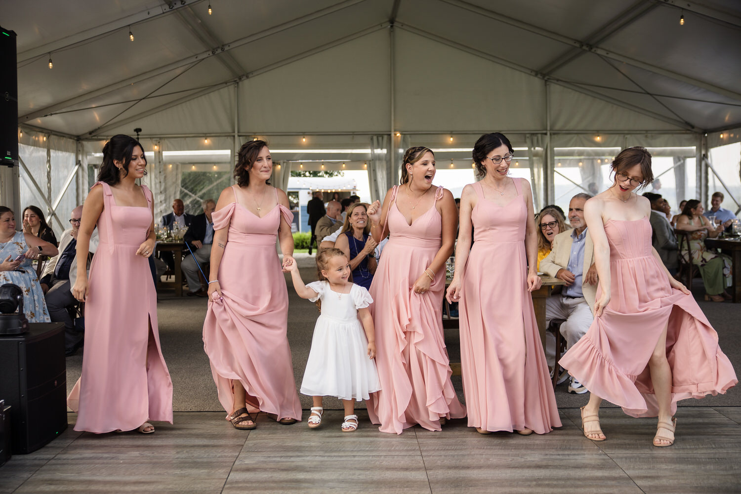 Five women in matching pink dresses and a young girl in white hold hands and smile while dancing together inside a large tent at a wedding reception. Guests watch from tables in the background.