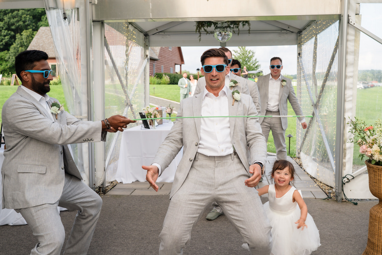 Three men in gray suits and sunglasses play limbo at an outdoor wedding, with a young girl in a white dress smiling nearby. The scene is festive and takes place under a decorated tent.