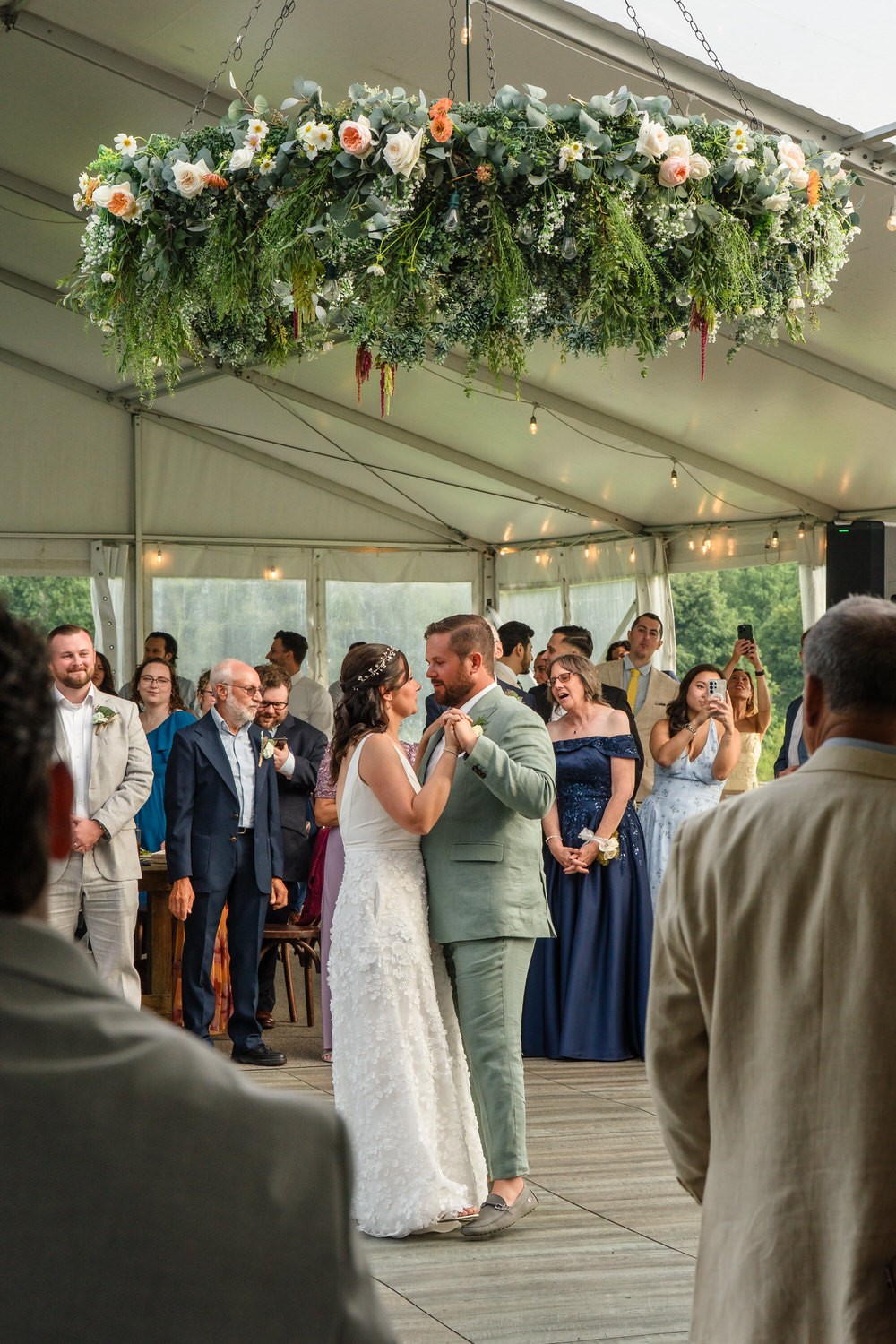 A bride and groom share their first dance under a floral chandelier at an outdoor wedding reception, surrounded by smiling guests who watch and take photos.