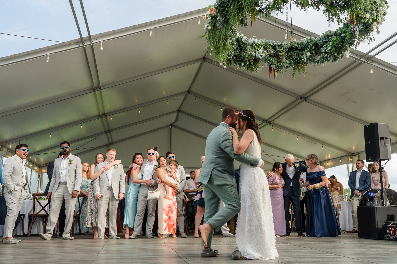 A bride and groom share a kiss while dancing at their wedding reception. Guests in formal attire watch and smile under a decorated, tented venue with greenery overhead.