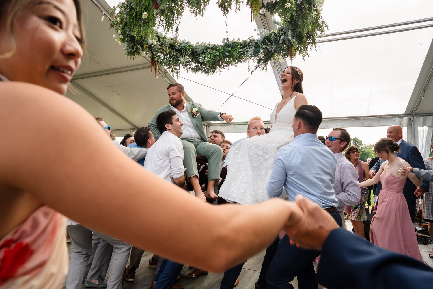 A bride and groom are lifted on chairs during a joyful wedding celebration, surrounded by guests holding hands and cheering under a floral canopy in a bright tent.