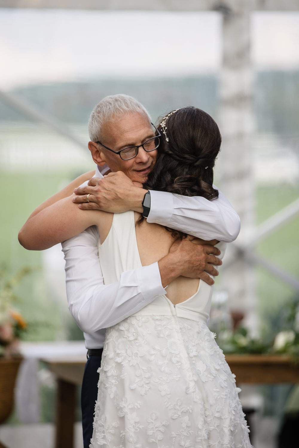 An older man with glasses hugs a woman in a white wedding dress, her back to the camera, in a joyful moment at a wedding celebration.