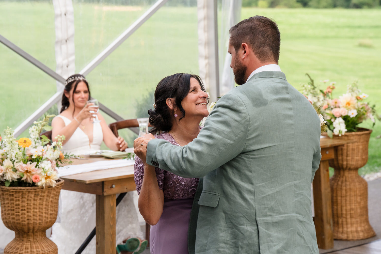 A man in a light green suit dances with a smiling woman in a lavender dress, while a bride sits at a table in the background, raising a glass. The setting is decorated with flowers and overlooks a green field.