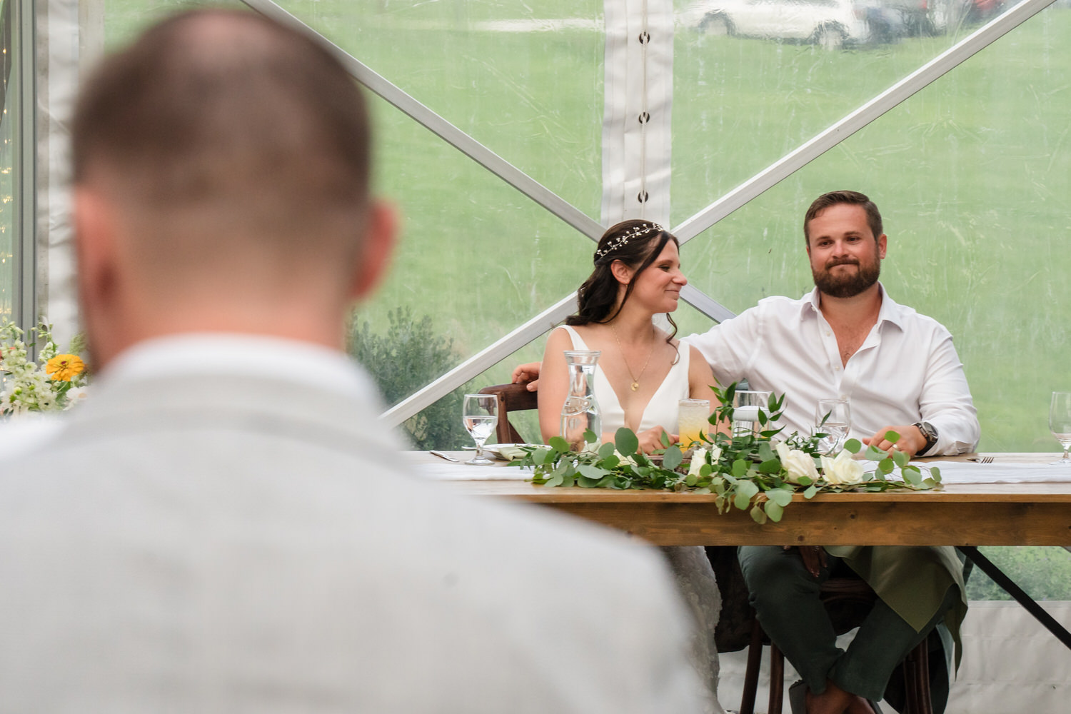A bride and groom sit together at a decorated table, smiling and looking toward a blurred figure in the foreground, inside a tent with a grassy background.