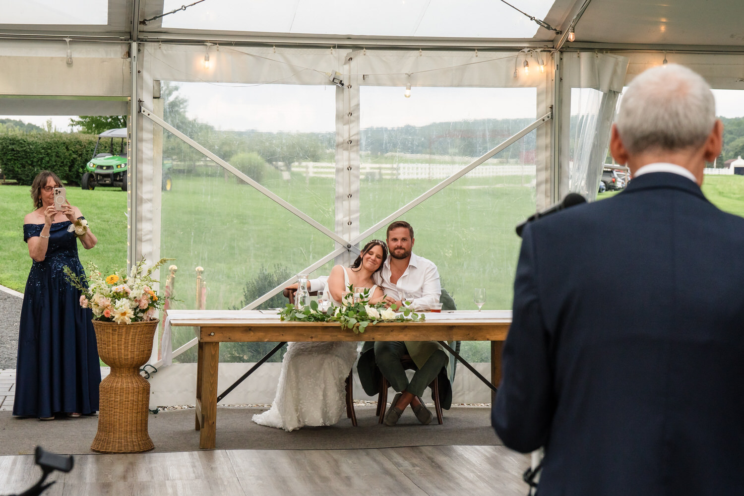 A bride and groom sit at a table under a tent, smiling and leaning into each other. An older man stands in the foreground speaking, while a woman in a blue dress takes a photo. Lush green lawn is visible outside.