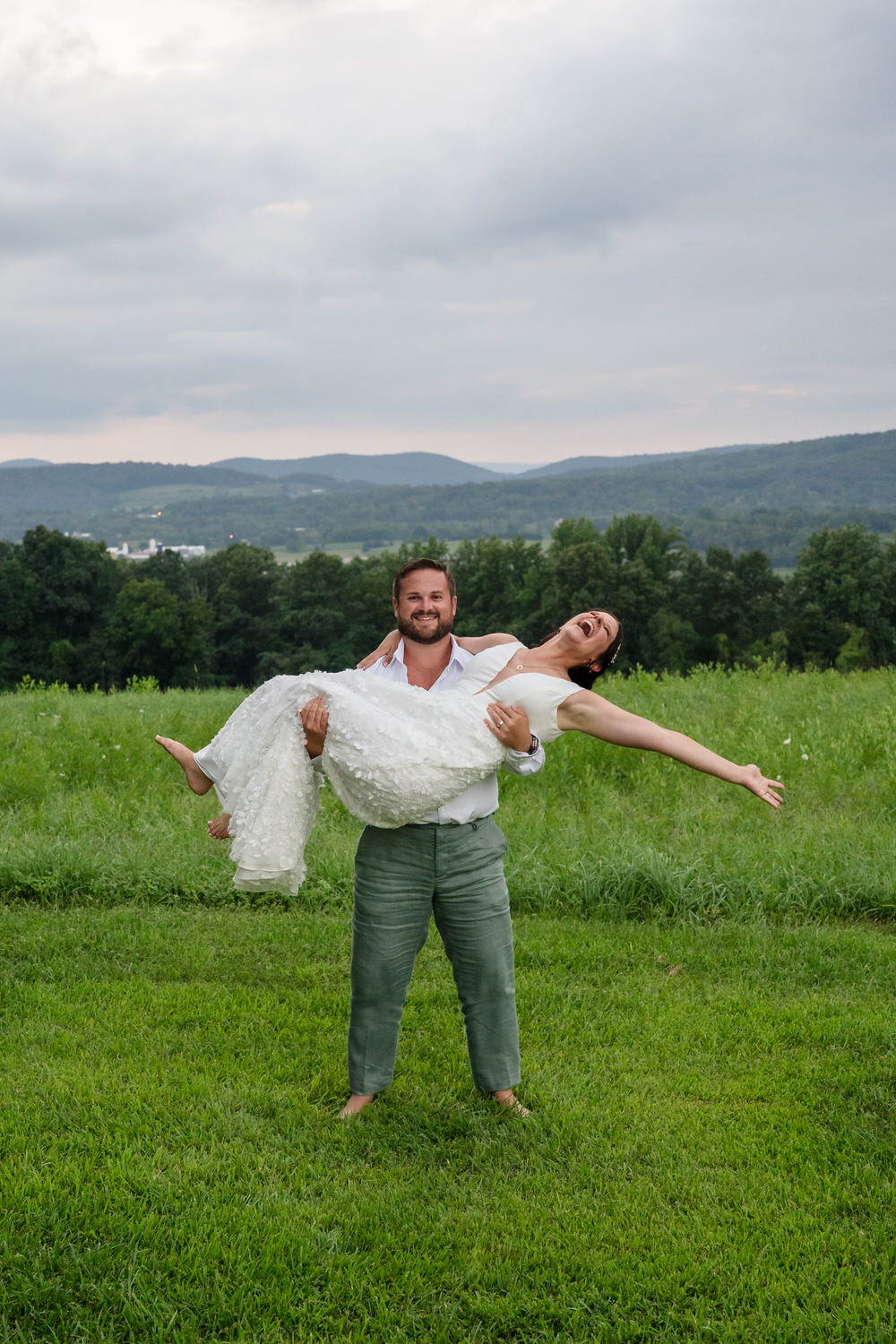 A man stands barefoot on grass, smiling as he lifts a laughing woman in a white dress. Behind them are green fields, trees, and rolling hills under a cloudy sky.