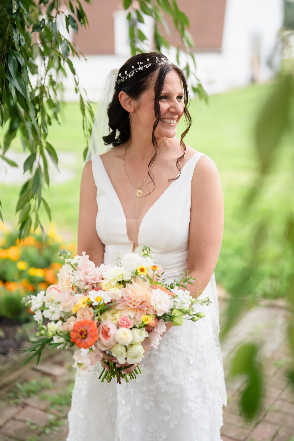 A smiling bride in a white dress holds a colorful bouquet of flowers outdoors, with greenery and yellow flowers visible in the background.