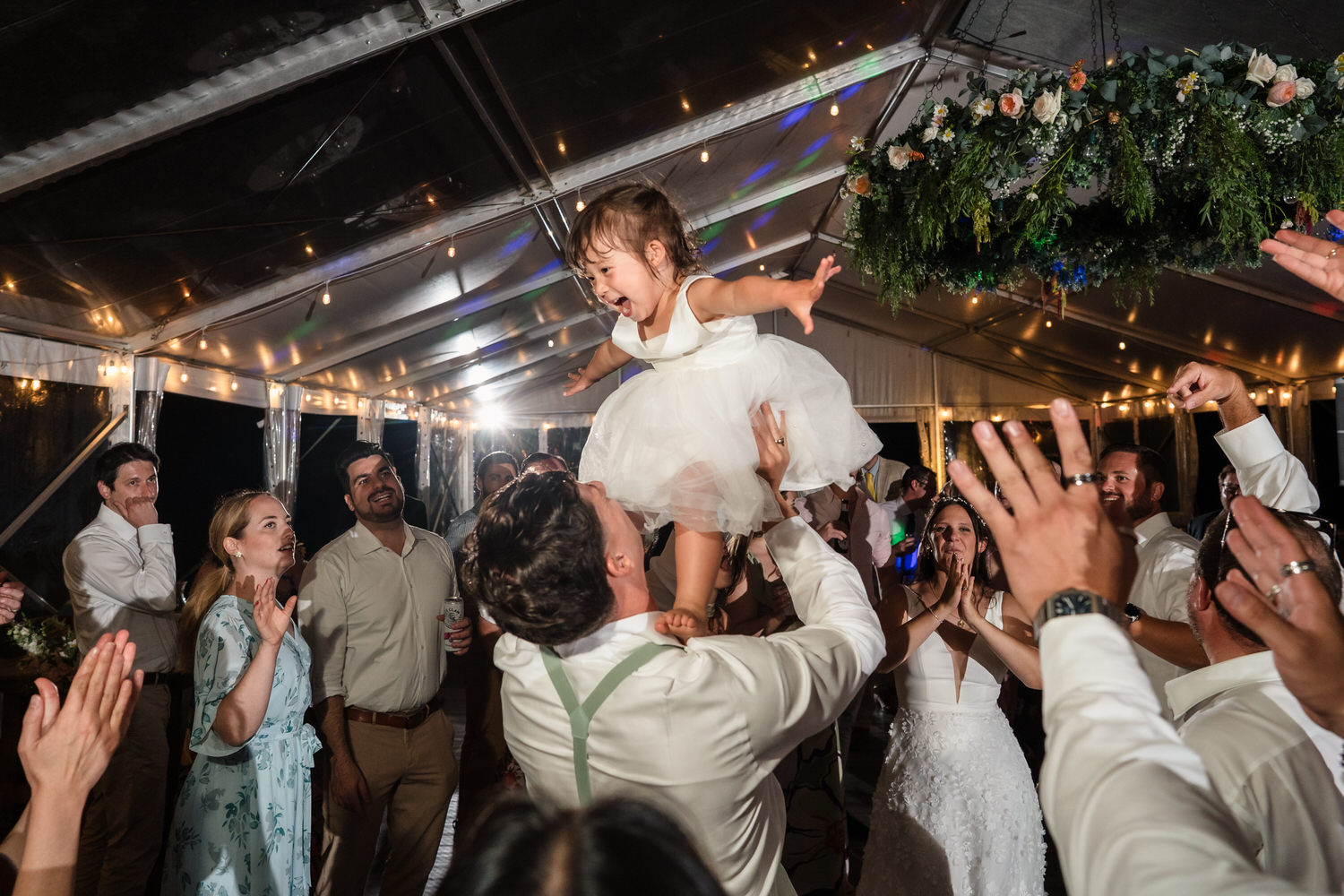 A man lifts a laughing young girl in a white dress at a lively wedding reception, surrounded by dancing guests under string lights and floral decorations.