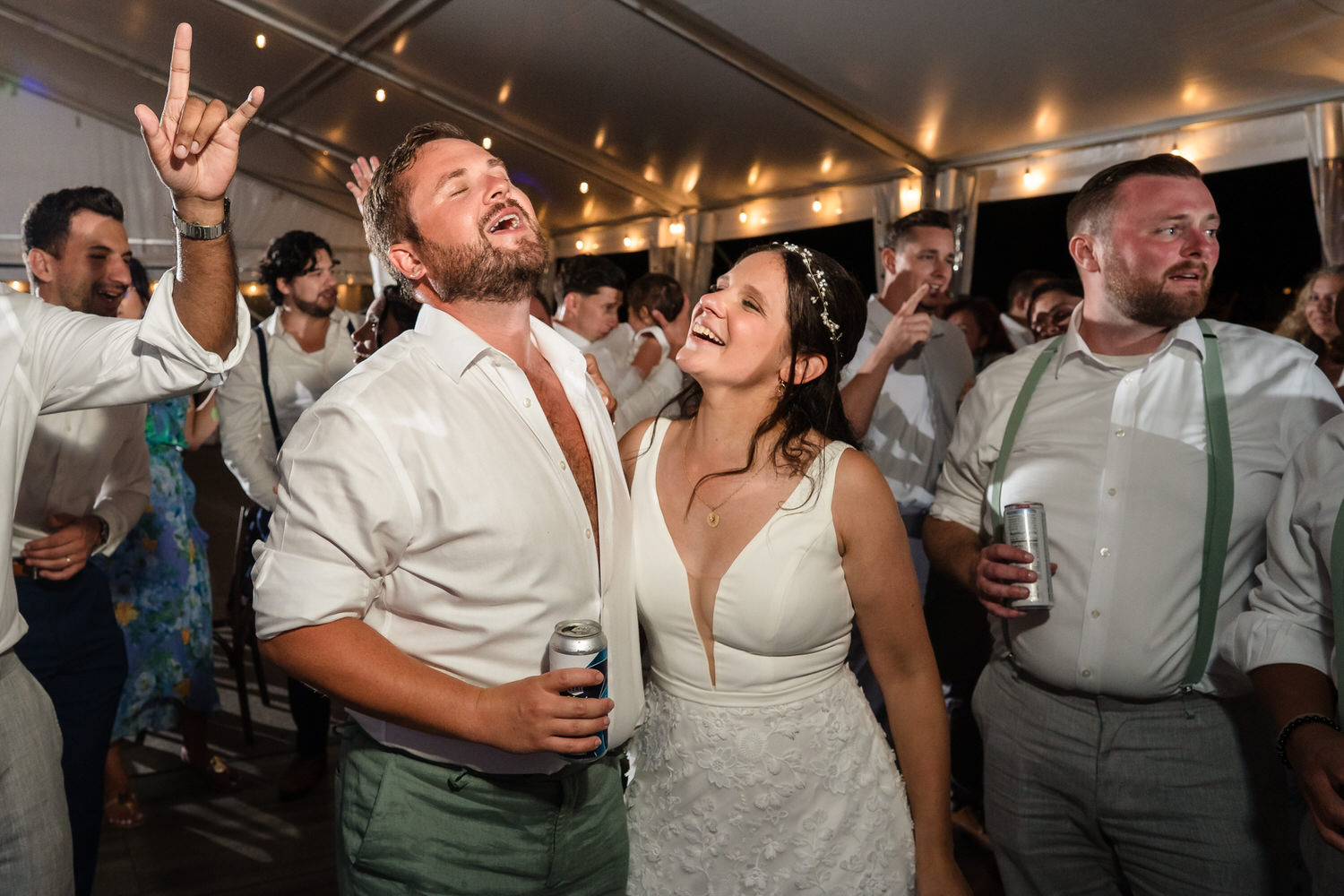 A bride and groom smile and celebrate on a dance floor, holding drinks. Guests surround them under string lights in a festive indoor setting.