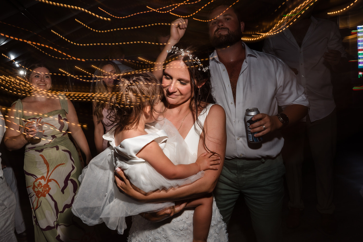A woman in a white dress holds a little girl while dancing at a party, surrounded by people and swirling string lights in the background.