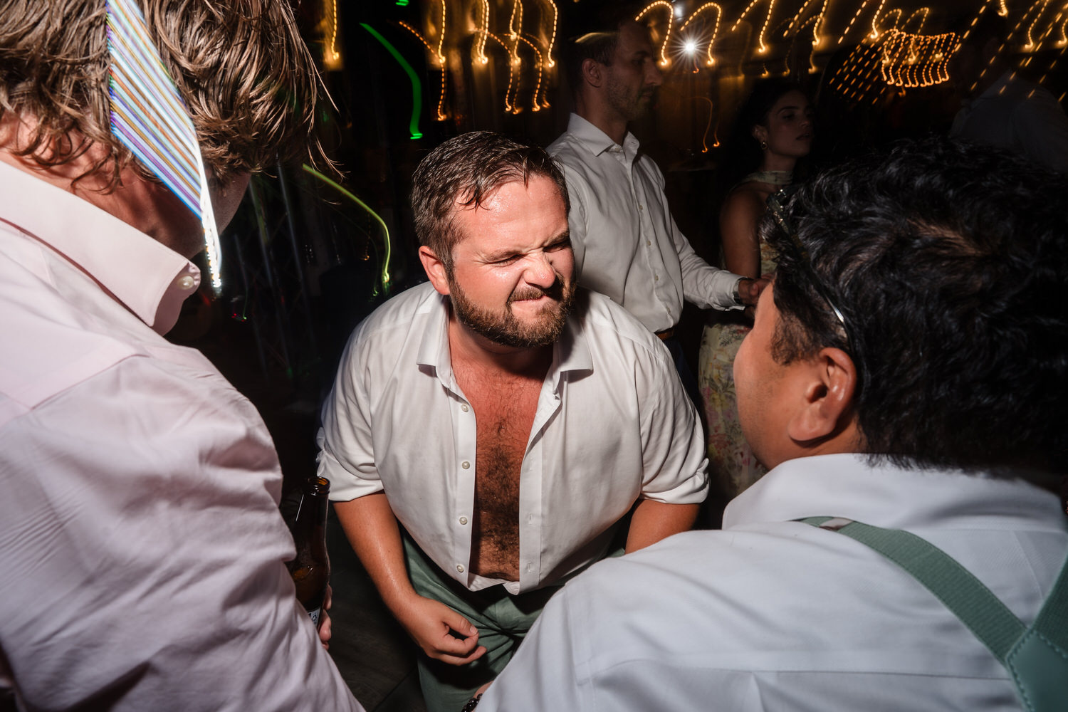 A man in an open white shirt makes a funny face while dancing with friends at a lively party, with bright string lights blurred in the background.