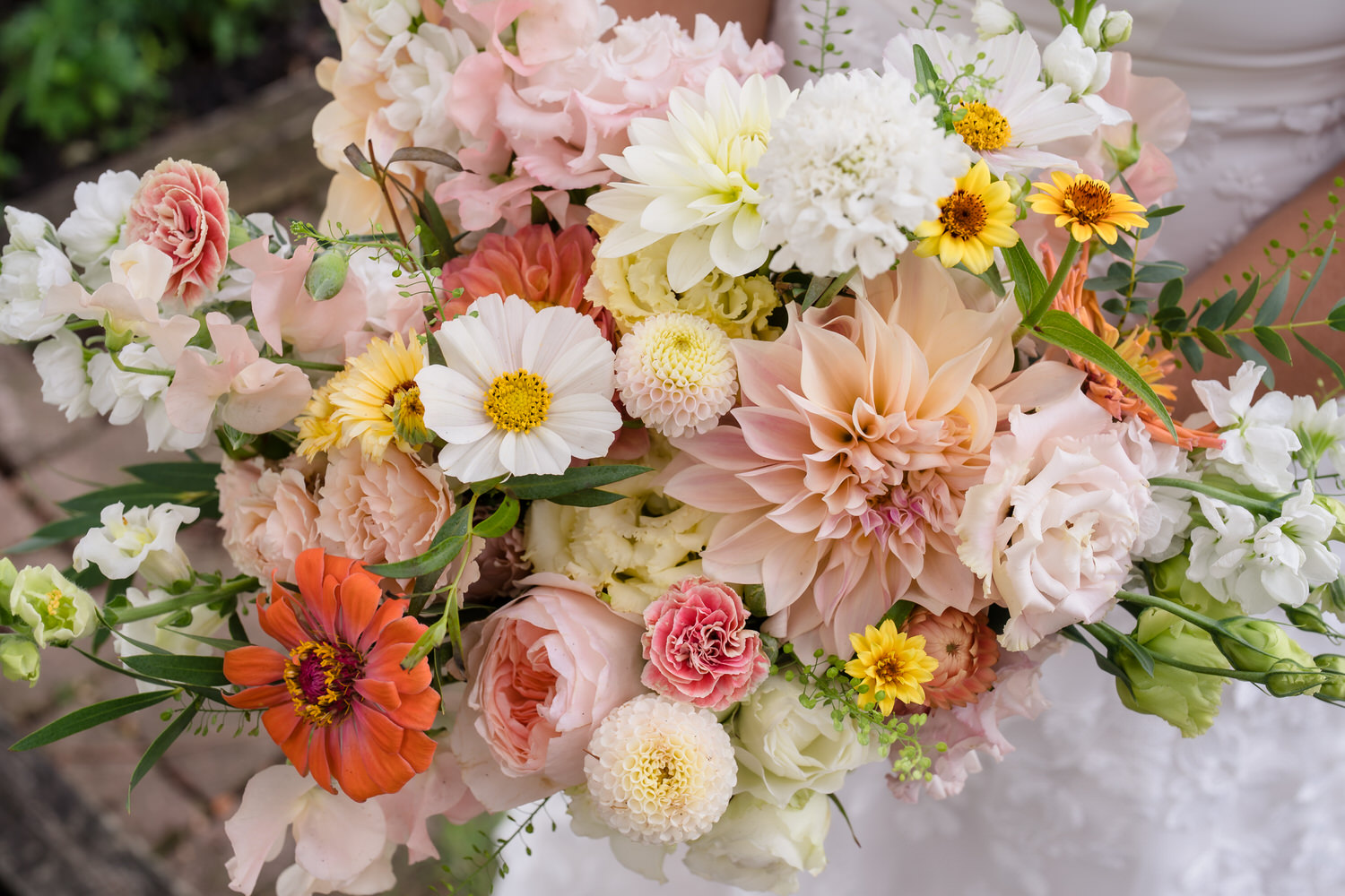 A close-up of a colorful bouquet featuring dahlias, roses, zinnias, and other assorted flowers in shades of pink, white, orange, and yellow, held by someone in a white dress.