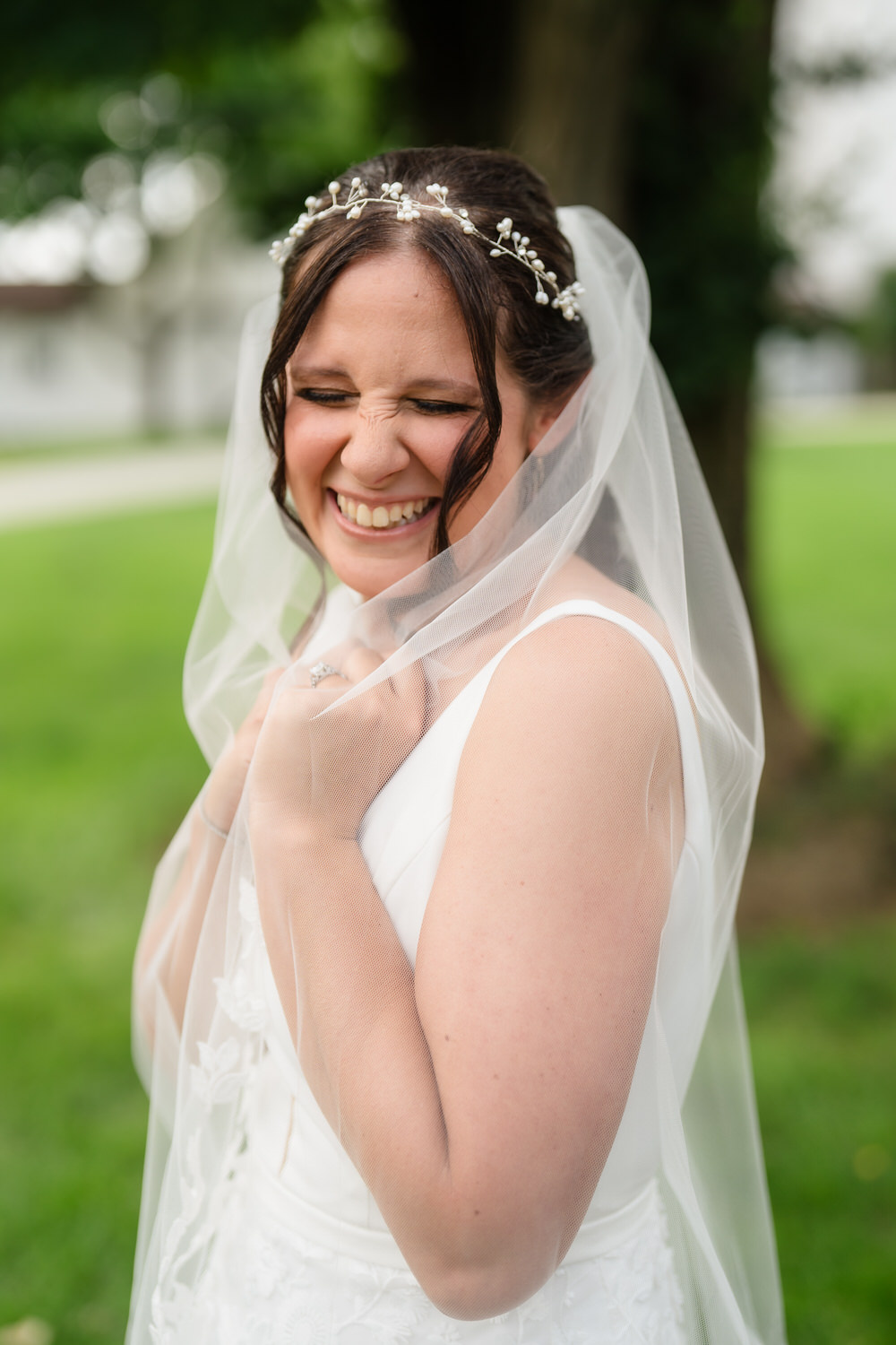 A smiling bride in a white dress and veil stands outdoors, eyes closed and gripping her veil joyfully, with greenery and a tree in the background.