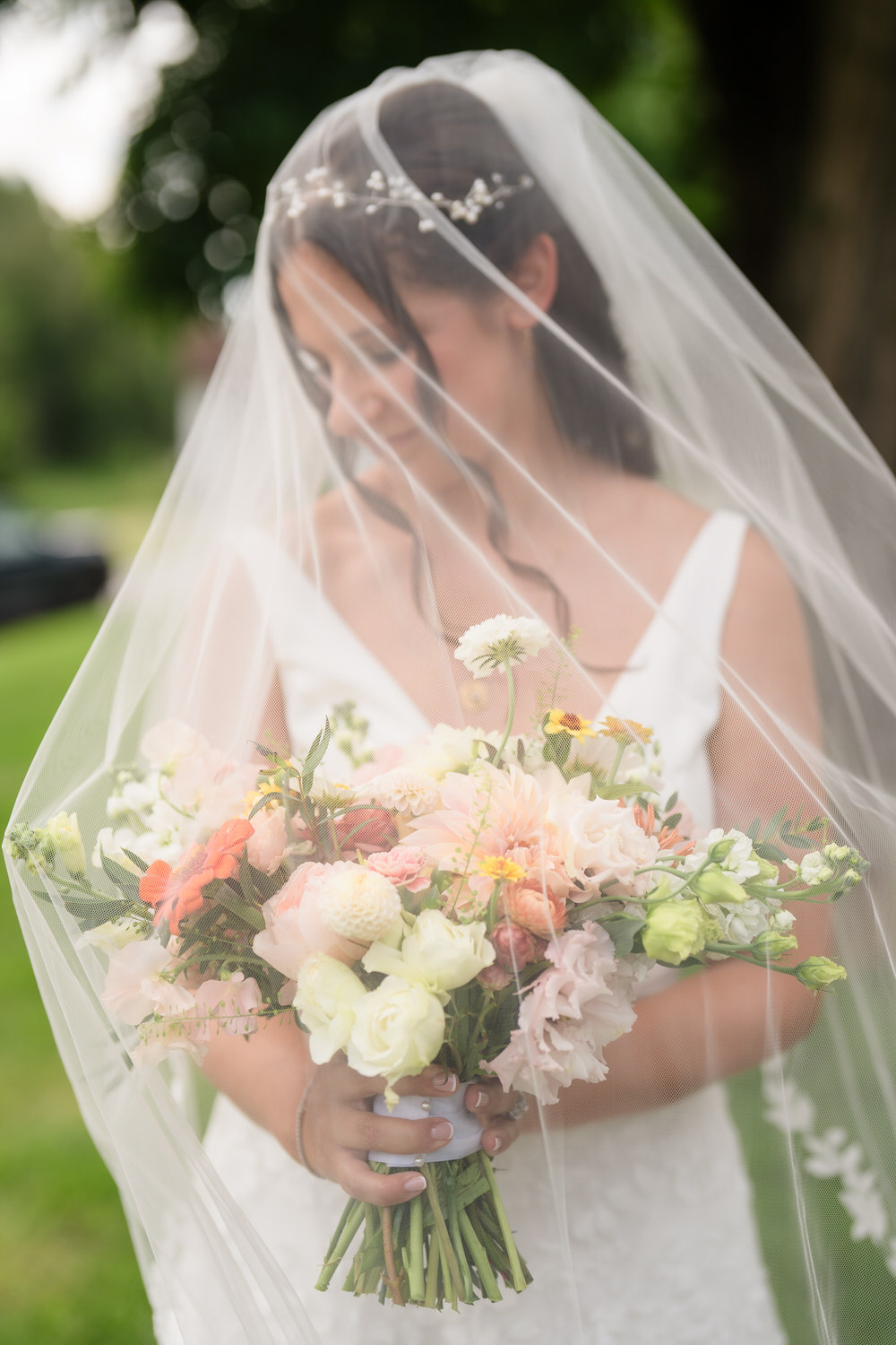 A bride in a white dress and veil holds a colorful bouquet of flowers, standing outdoors on a green lawn, with her face softly obscured by the veil.
