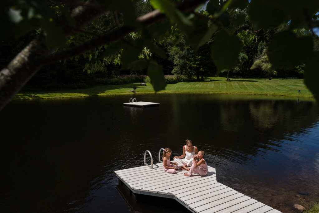 A woman and two children sit on a small dock over a pond, surrounded by trees and grass, with a floating platform visible in the water nearby—a peaceful moment reminiscent of Farm Road Estate wedding photos, capturing the beauty and tranquility of countryside settings.