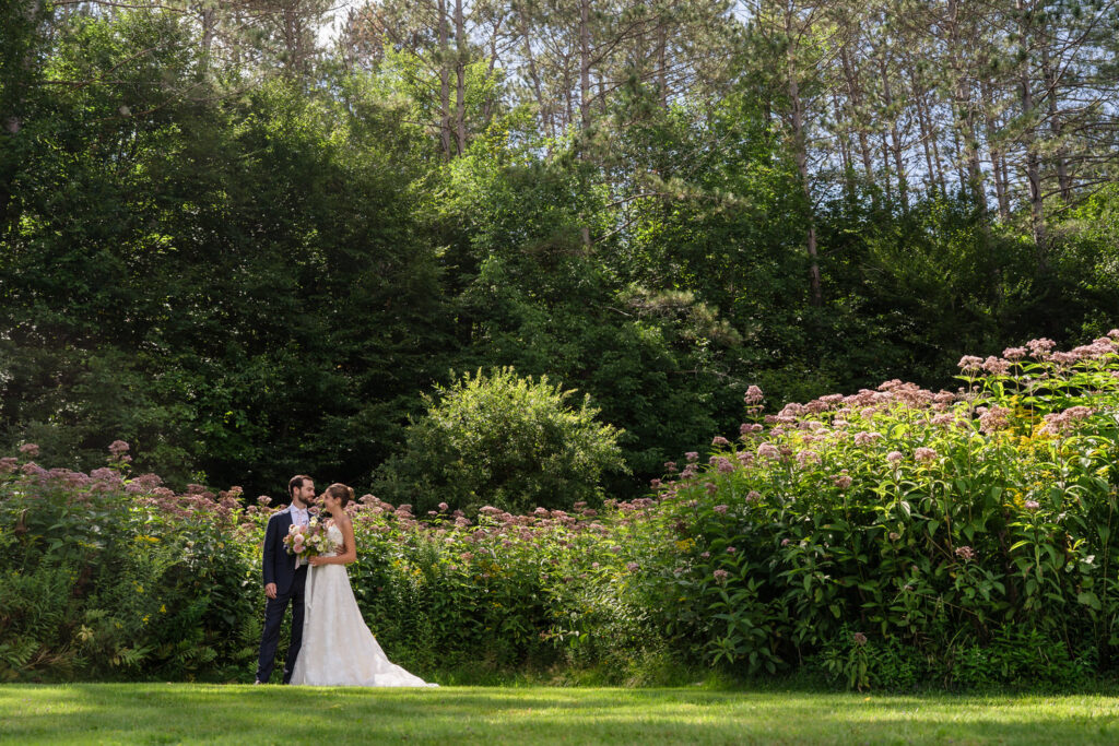 A bride and groom stand close together in a lush garden surrounded by tall flowering plants, beneath a canopy of trees and a bright blue sky, capturing the romantic essence of Farm Road Estate wedding photos.
