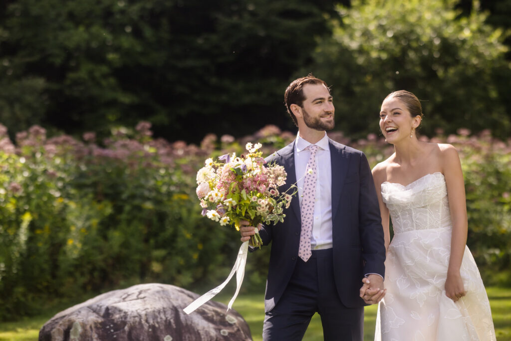 A couple dressed in wedding attire holds hands and smiles outdoors at their Farm Road Estate wedding; the groom holds a large bouquet of flowers. They stand on grass with lush greenery and pink flowers in the background, capturing the charm of Farm Road Estate wedding photos.