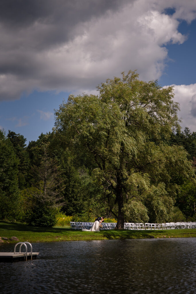 A couple stands beneath a large tree beside a calm lake, with rows of white chairs arranged on the green grass in the background under a partly cloudy sky, capturing the serene beauty often seen in Farm Road Estate wedding photos.