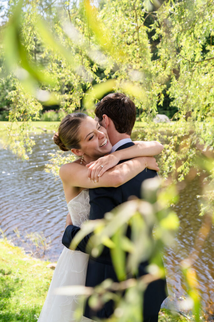 A bride and groom embrace joyfully by a sunlit pond at Farm Road Estate, surrounded by green leaves and trees, with the bride smiling brightly and wearing a white dress.
