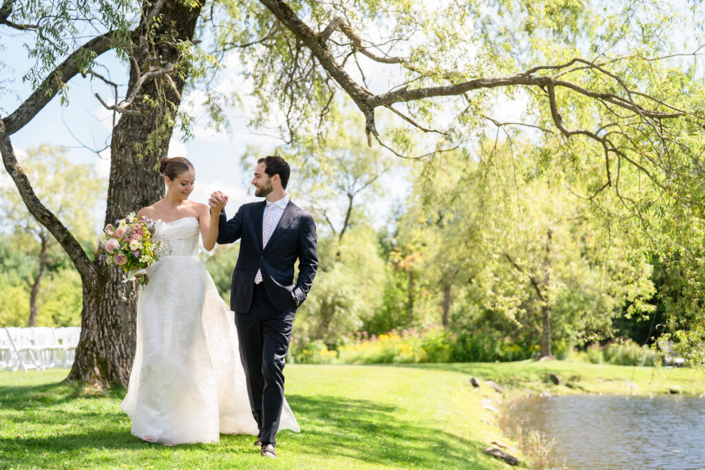 A bride in a white gown and a groom in a dark suit walk hand-in-hand on a sunny lawn by a pond at Farm Road Estate, surrounded by green trees and flowers, smiling at each other—the perfect moment captured in timeless Farm Road Estate wedding photos.