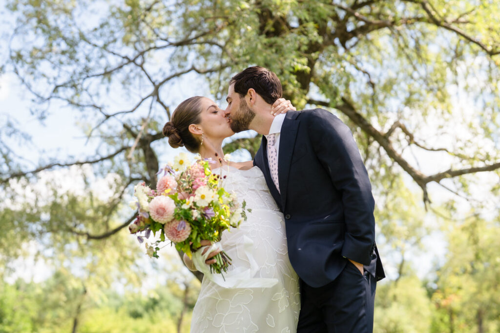 A bride and groom share a kiss outdoors in this stunning Farm Road Estate wedding photo. The bride holds a bouquet of flowers and wears a white dress, while the groom wears a navy suit. Green trees and sunlight create a romantic, natural background.