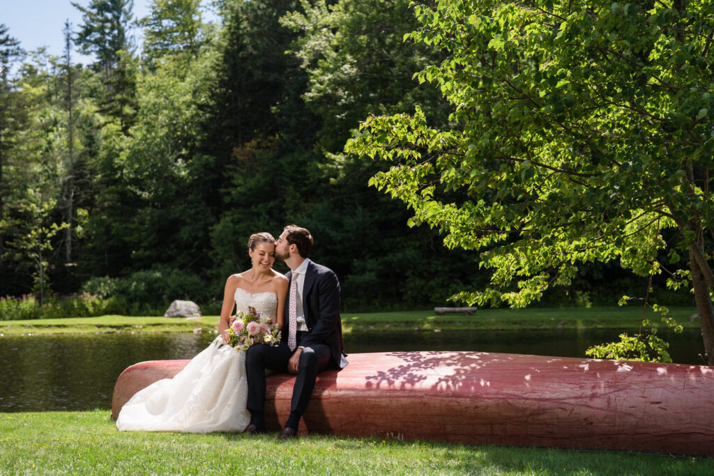 A bride and groom sit on a large red canoe by a pond at Farm Road Estate, surrounded by lush green trees on a sunny day. The bride holds a bouquet and smiles while the groom kisses her temple, creating the perfect moment for Farm Road Estate wedding photos.
