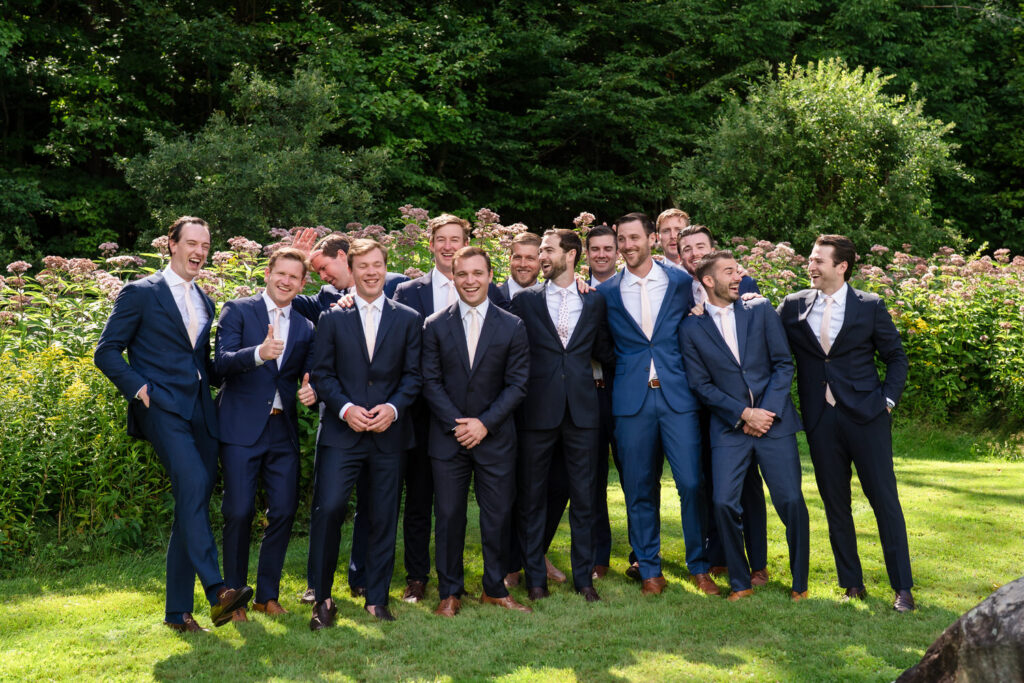 Twelve men in suits stand outdoors on grass, smiling and laughing together in front of green bushes and trees on a sunny day, capturing joyful moments for Farm Road Estate wedding photos.