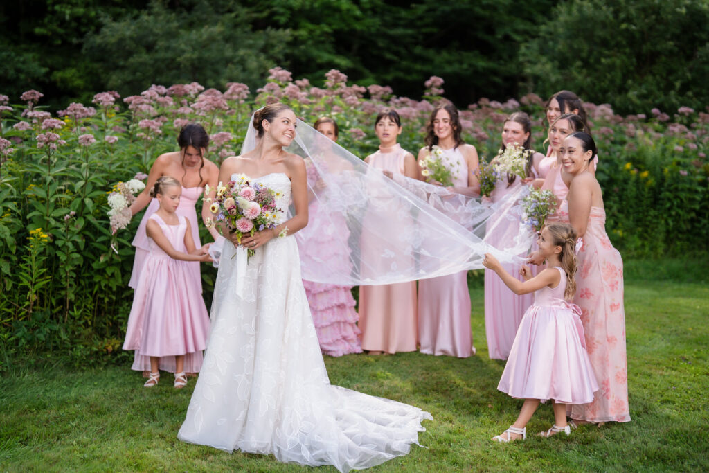 A bride in a white dress stands outside with bridesmaids and flower girls in pink dresses. The group smiles and holds her veil in a garden setting with tall flowers and greenery, capturing the joy of Farm Road Estate wedding photos.