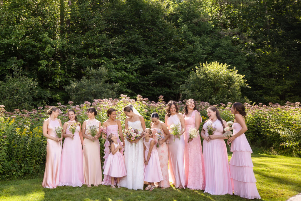 Eleven women and two girls in pale pink dresses stand outside in a garden at Farm Road Estate, holding bouquets and smiling. Trees and flowering bushes are in the background, and sunlight filters through the scene, capturing the perfect wedding photo moment.