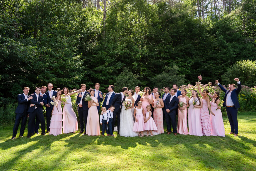 A wedding party poses outdoors for beautiful Farm Road Estate wedding photos; people in pink dresses and dark suits stand on grass, smiling and cheering, with trees in the background.