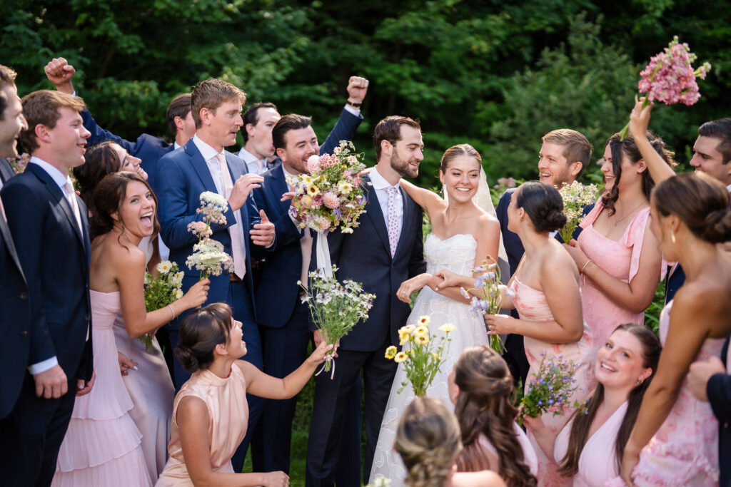 A bride and groom smile surrounded by a joyful wedding party outdoors in this beautiful Farm Road Estate Wedding photo. Bridesmaids in pink dresses hold flowers, while groomsmen in suits cheer and celebrate, with lush greenery in the background.