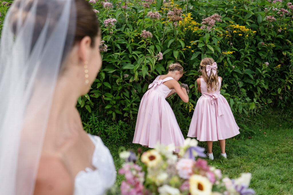 A bride in a veil holds a bouquet in the foreground, while two girls in pink dresses stand by tall flowers along the Farm Road Estate, picking or examining something with their backs to the camera—capturing timeless wedding photos.
