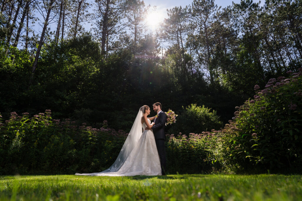 A bride and groom stand together outdoors in a sunlit garden at Farm Road Estate, surrounded by tall trees and greenery, gazing at each other while holding a bouquet—perfectly capturing the essence of Farm Road Estate wedding photos.