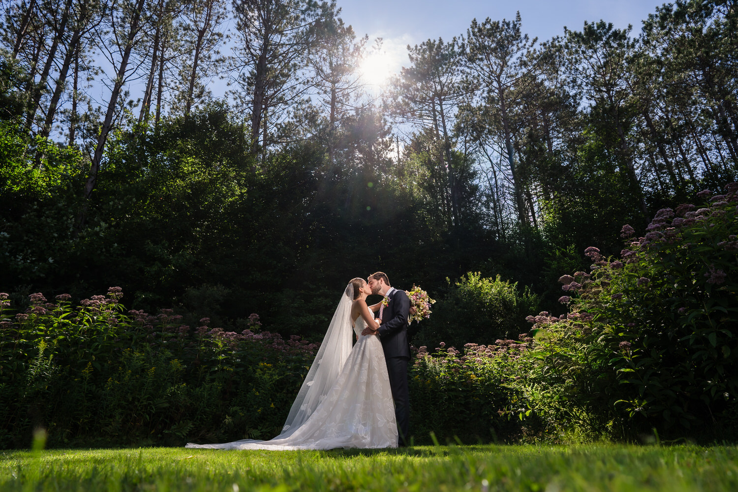A bride and groom embrace and kiss in a lush garden at Farm Road Estate, surrounded by tall trees and flowers, with sunlight streaming through the trees above them—capturing the timeless romance of Farm Road Estate wedding photos.