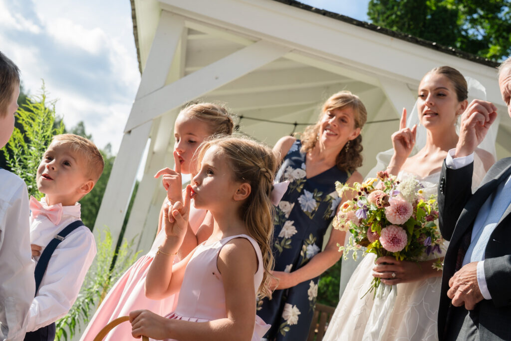 A bride, an older woman, and children in pastel clothes are outdoors near a white gazebo at Farm Road Estate, holding up fingers and smiling, with a bouquet of flowers in the bride’s hand—capturing the joy of timeless Farm Road Estate wedding photos.