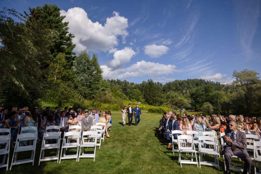 A wedding ceremony outdoors on a sunny day; guests sit on white chairs while a couple walks down the aisle, surrounded by trees and a blue sky with clouds—capturing the timeless romance found in Farm Road Estate wedding photos.