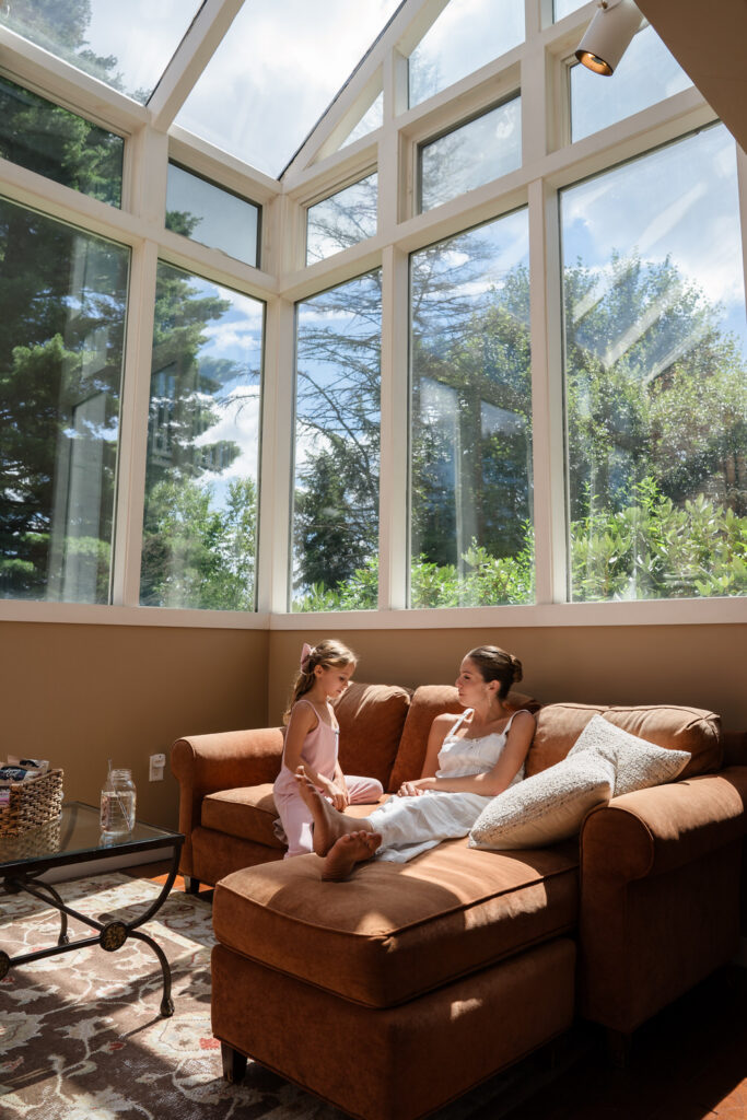 A woman relaxes on a brown sectional sofa while a young girl stands beside her in a sunlit room with large windows and views of trees outside, reminiscent of the serene atmosphere often captured in Farm Road Estate wedding photos.