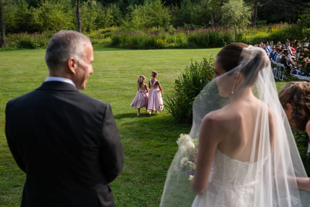 A bride and man watch two young girls in pink dresses walking across a grassy lawn toward them at an outdoor wedding ceremony at Farm Road Estate, with guests seated in the background—capturing a timeless moment among their Farm Road Estate Wedding Photos.