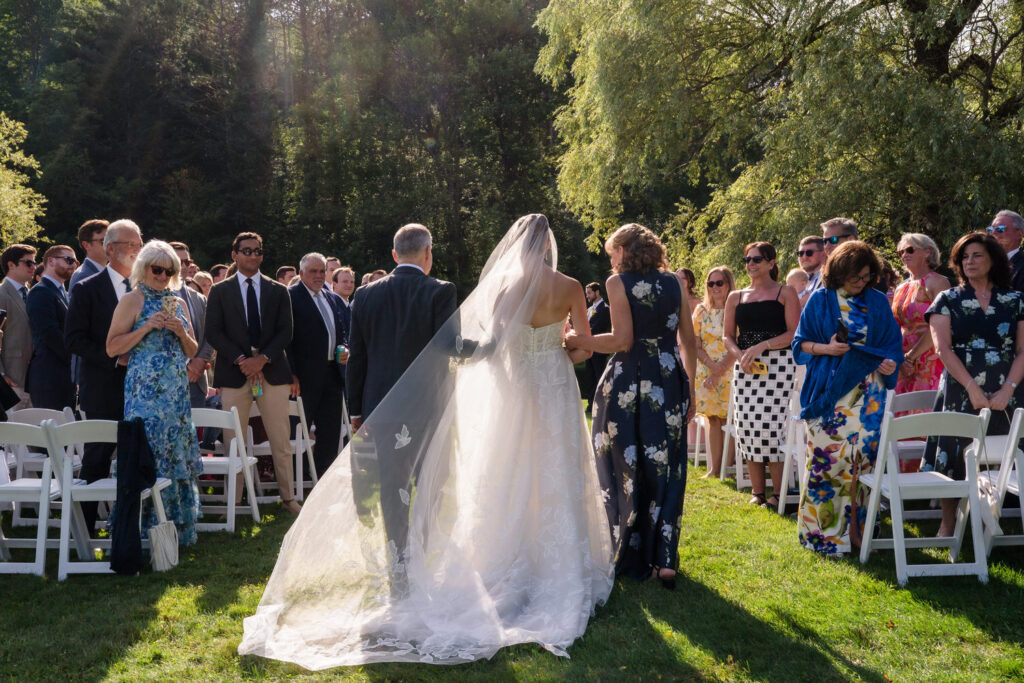 A bride in a white gown walks down the aisle outdoors at Farm Road Estate, escorted by two people, with guests seated and standing on either side, all dressed in formal attire on a sunny day—capturing the essence of timeless Farm Road Estate wedding photos.