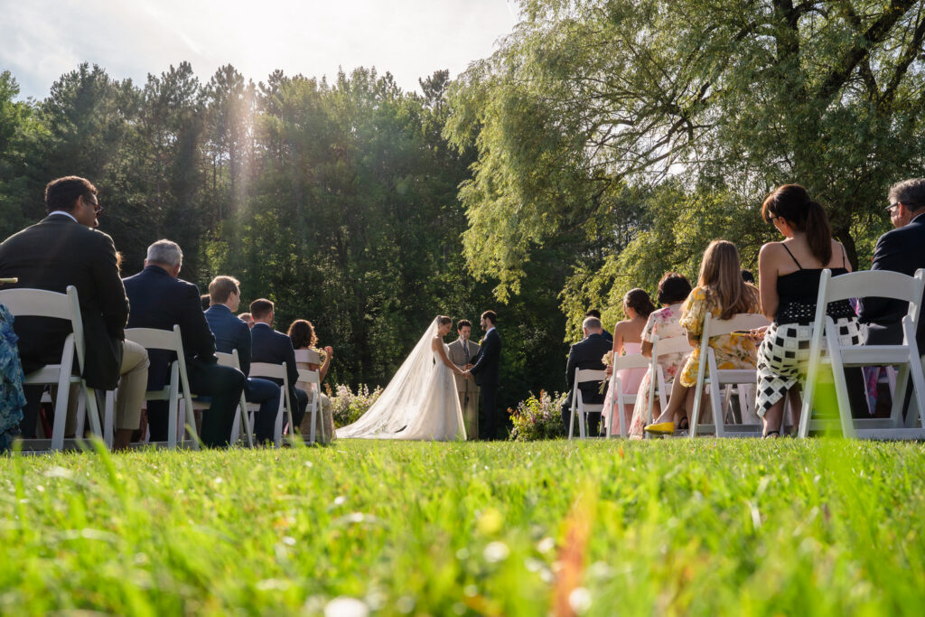 A bride and groom stand facing each other outdoors during their wedding ceremony at Farm Road Estate, surrounded by seated guests on white chairs, with sunlight streaming through trees in the background—capturing the essence of unforgettable Farm Road Estate wedding photos.