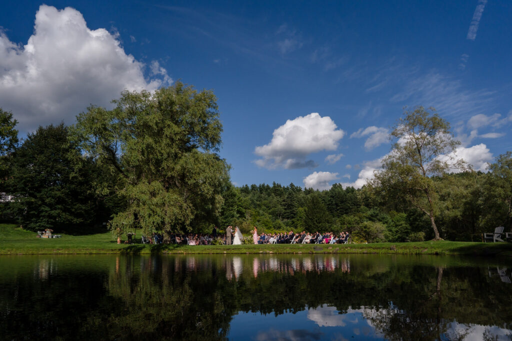 A wedding ceremony takes place outdoors beside a pond at Farm Road Estate, surrounded by trees and greenery, with guests seated under a blue sky with fluffy clouds reflected in the water—creating picture-perfect Farm Road Estate wedding photos.