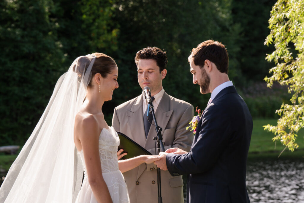 A bride and groom stand outdoors exchanging rings during their Farm Road Estate Wedding Ceremony as an officiant reads from a folder. Trees and a pond are in the background, capturing the natural beauty showcased in Farm Road Estate Wedding Photos.