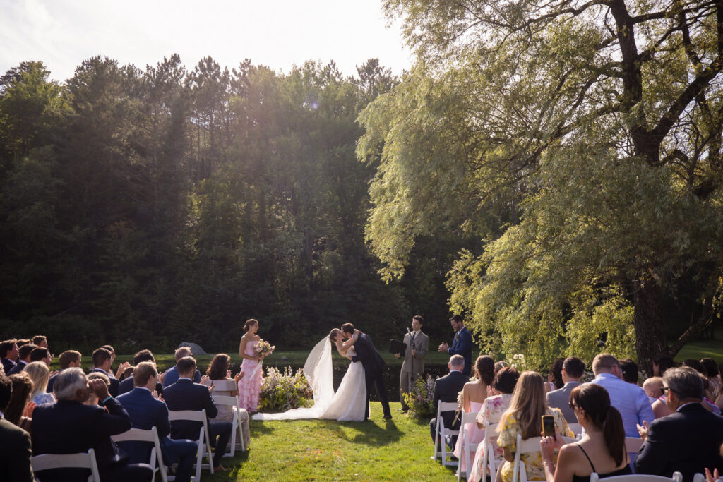 A couple kisses at an outdoor Farm Road Estate wedding ceremony, surrounded by guests seated in white chairs, lush greenery, and tall trees in the background under sunlight.