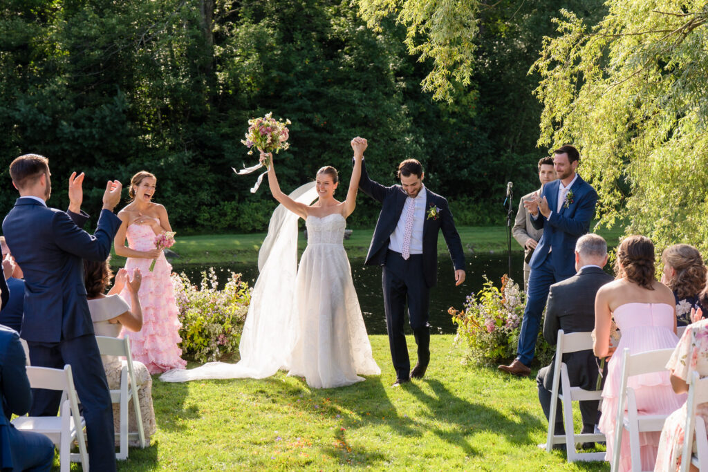 A bride and groom celebrate at an outdoor Farm Road Estate wedding ceremony, holding hands and raising arms as guests seated on white chairs clap and cheer. Green trees and a small pond are in the background, perfectly capturing the joy in these Farm Road Estate wedding photos.