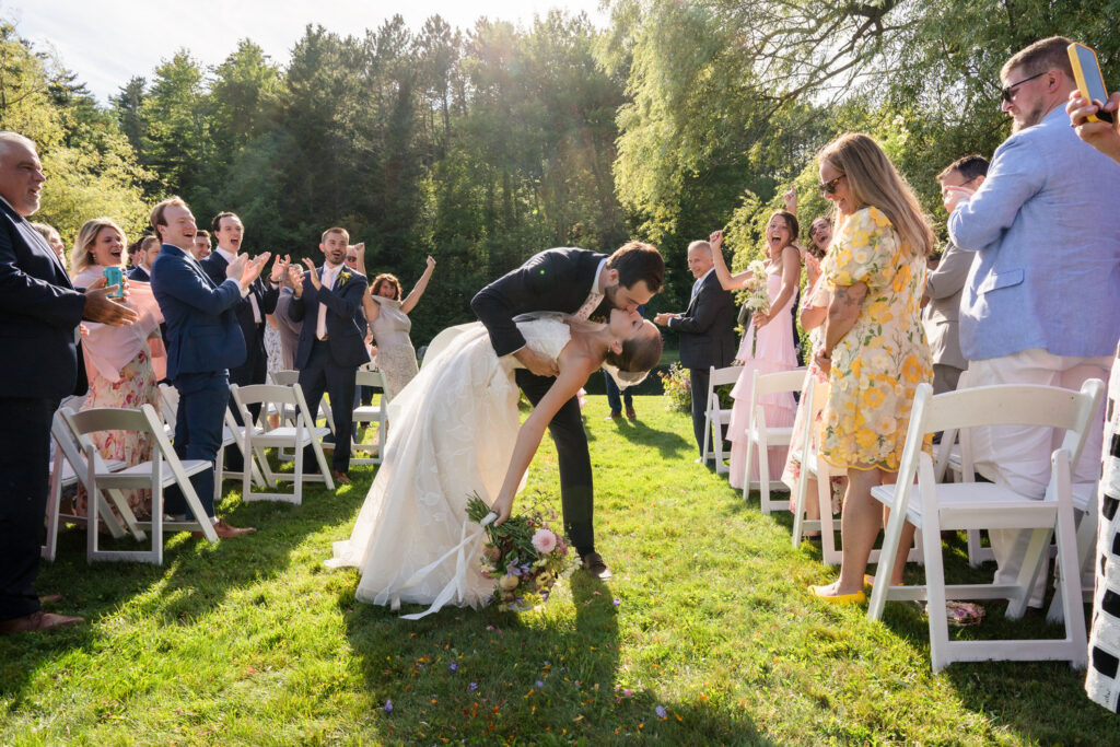 A newlywed couple kisses as the groom dips the bride in the aisle, surrounded by clapping and cheering guests outdoors on a sunny day—capturing one of many joyful Farm Road Estate wedding photos.