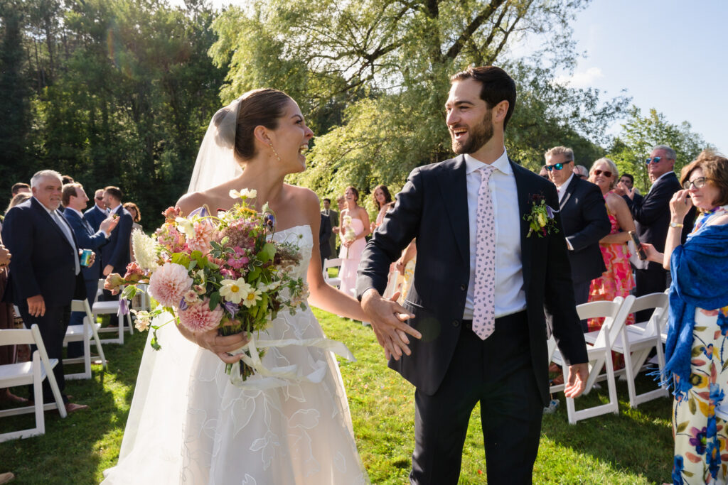 A bride and groom smile joyfully as they walk hand in hand down the aisle outdoors at Farm Road Estate, surrounded by applauding guests on a sunny day. The bride holds a colorful bouquet of flowers, perfectly captured in these unforgettable Farm Road Estate wedding photos.