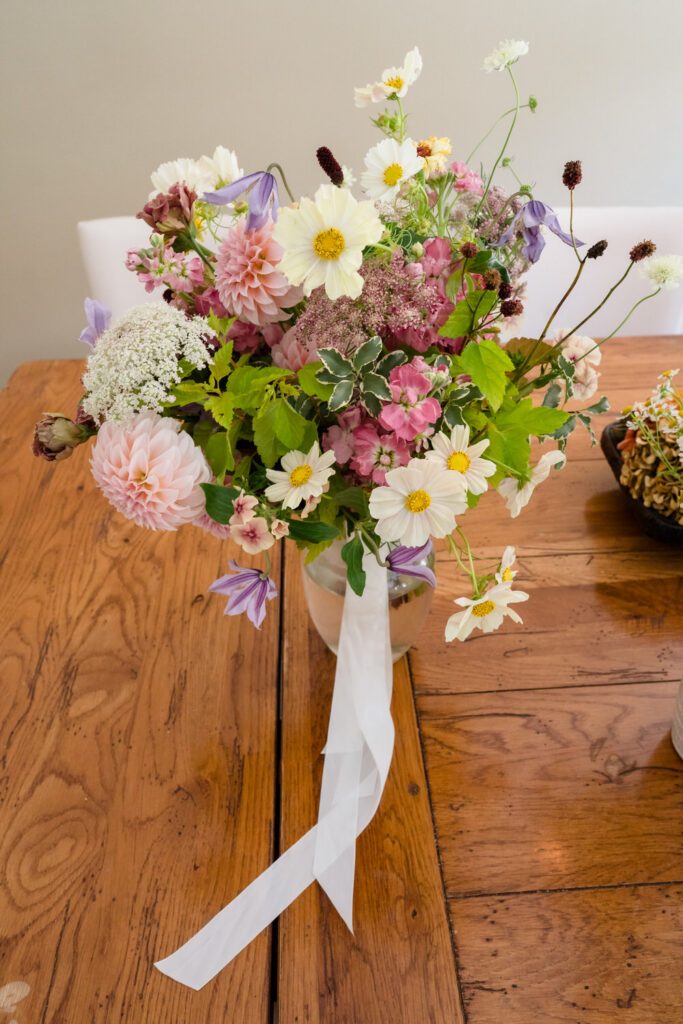A glass vase with a mixed bouquet of pink, white, and purple flowers with greenery and a white ribbon, placed on a wooden table—perfect for capturing elegant Farm Road Estate wedding photos.